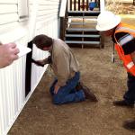 City housing inspector in orange vest and hardhat watches landlord paint the side of a rental unit exterior.