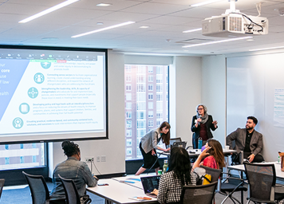 A diverse group of people in a modern conference room engages in a presentation. A large screen displays slides, and one person presents confidently.