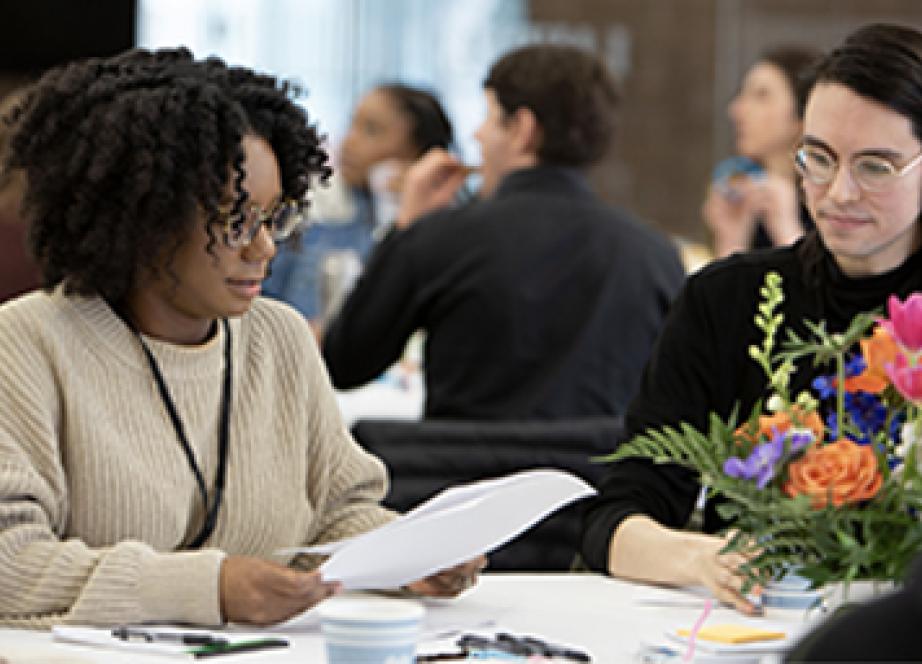 Two people sitting at a table reviewing papers; one wears a beige sweater, the other a black top. A vibrant bouquet is in the foreground, creating a focused, professional atmosphere.